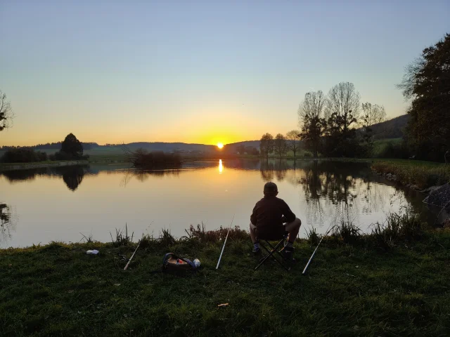Pêche sur le territoire. Un pêcheur avec trois cannes regardant le soleil se coucher à l'horizon. L'étang couvre la moitié du plan et fait refléter le paysage comme dans un miroir