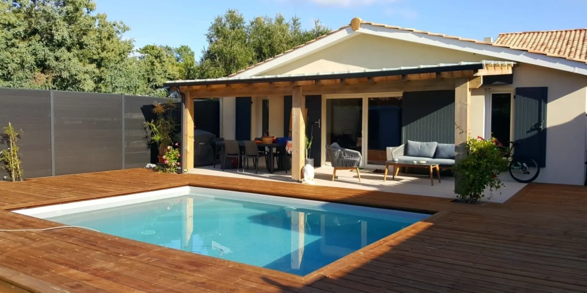Vue extérieure du meublé de tourisme L'Annexe avec sa piscine privée encastrée dans une grande terrasse en bois, un salon de jardin moderne sous une pergola et une façade de maison de vacances typique de Vendays-Montalivet.