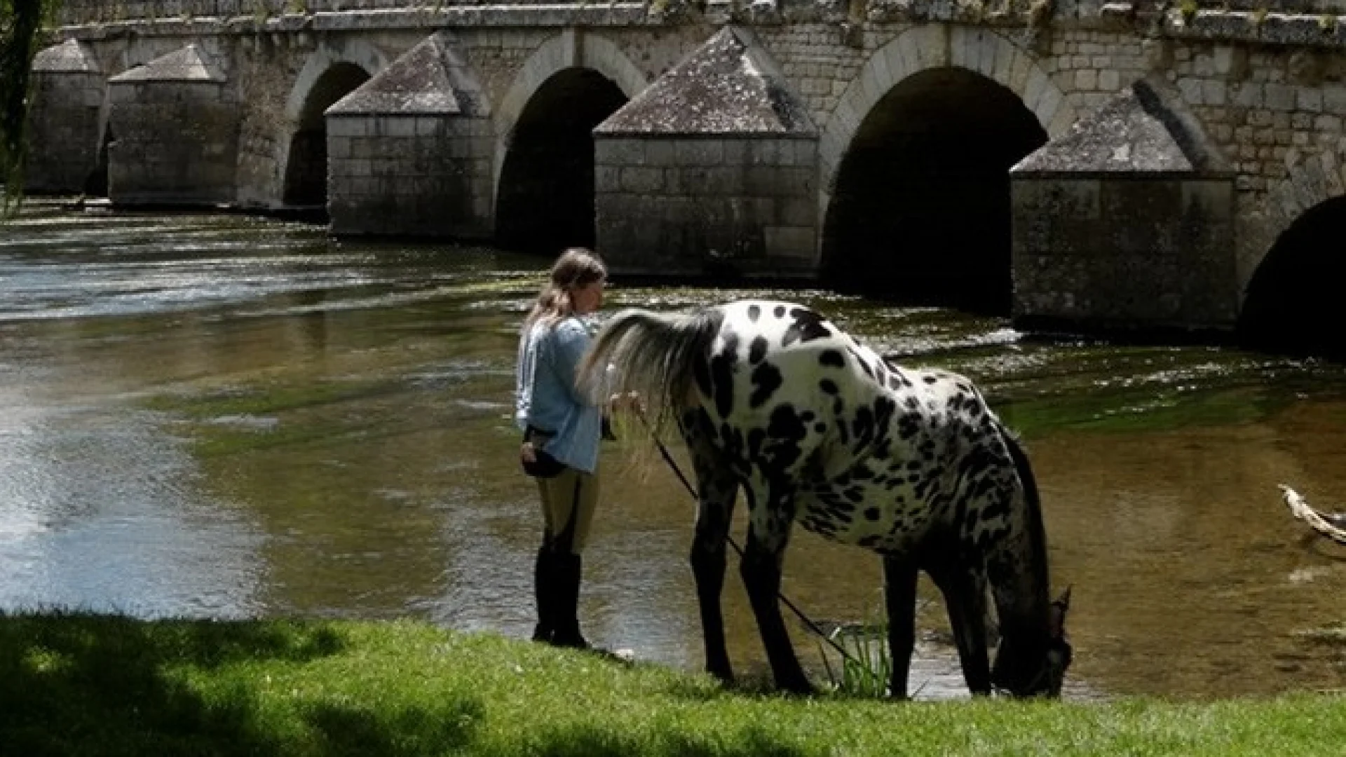 Cheval et sa cavalière au bord d'une rivière et d'un pont ancien avec les restes d'une forteresse en fond