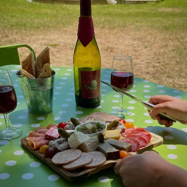 Table en bord en rivière avec bouteille de vin, verres... avec des mains et une planches de charcuterie et pain dans un seau