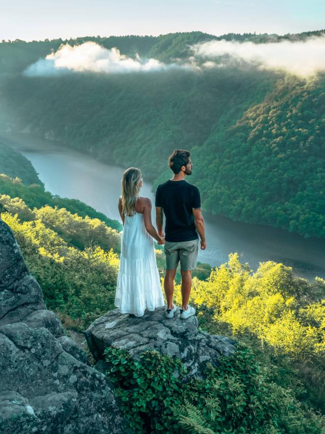 Une femme blonde avec une robe blanche et un homme brun en tee-shirt noir et short gris, se tiennent la main et regardent la rivière Dordogne et ses gorges depuis le point de vue du Roc du Busatier.