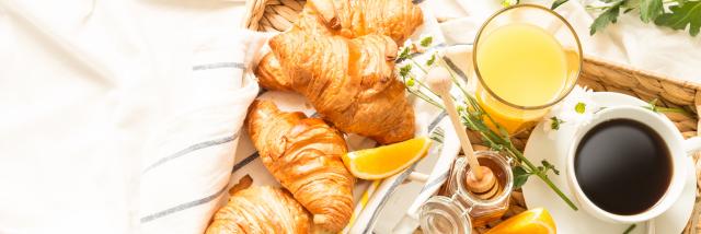 Continental breakfast on white bed sheets. Coffee, orange juice, croissants, jam, honey and flowers on wicker tray from above (top view, flat lay). Background layout with free text (copy) space.